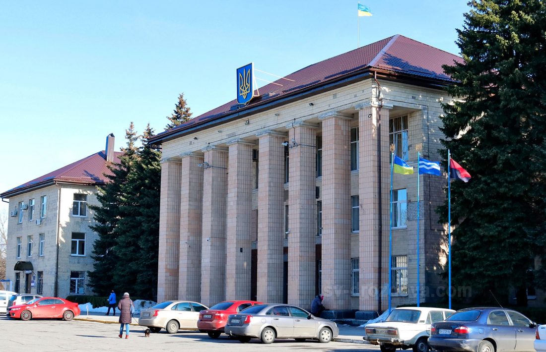Велика будівля з колонами та червоним дахом. На фасаді будівлі розміщено герб України, а на даху майорить український прапор. Перед будівлею стоять кілька автомобілів, а люди прогулюються поруч. На вулиці встановлені флагштоки з прапорами. Будівля оточена деревами.
