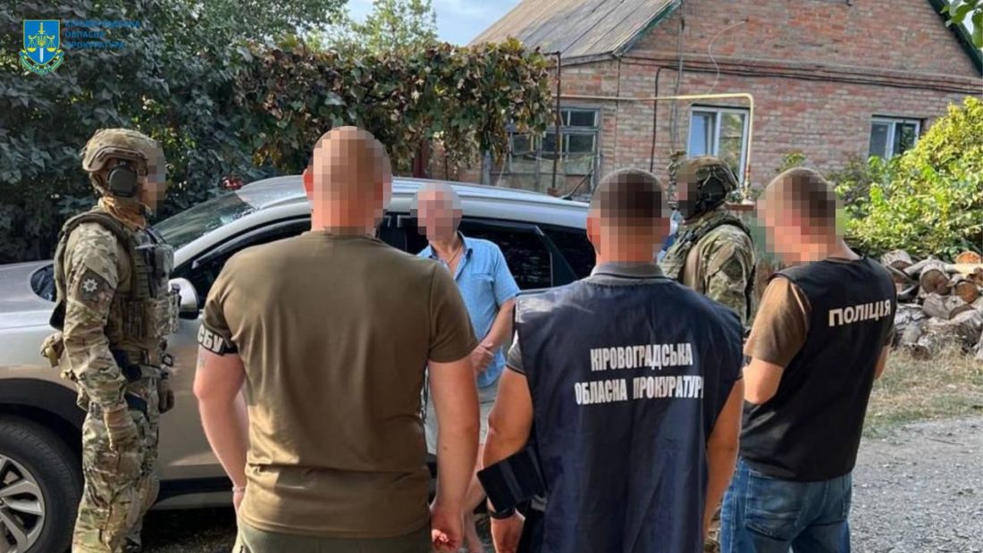 Група людей біля автомобіля. Двоє з них у формі з написами "Кіровоградська обласна прокуратура" і "Поліція", а інші одягнуті в камуфляжну форму. Їхні обличчя розмиті. На задньому плані видно цегляний будинок і рослинність.