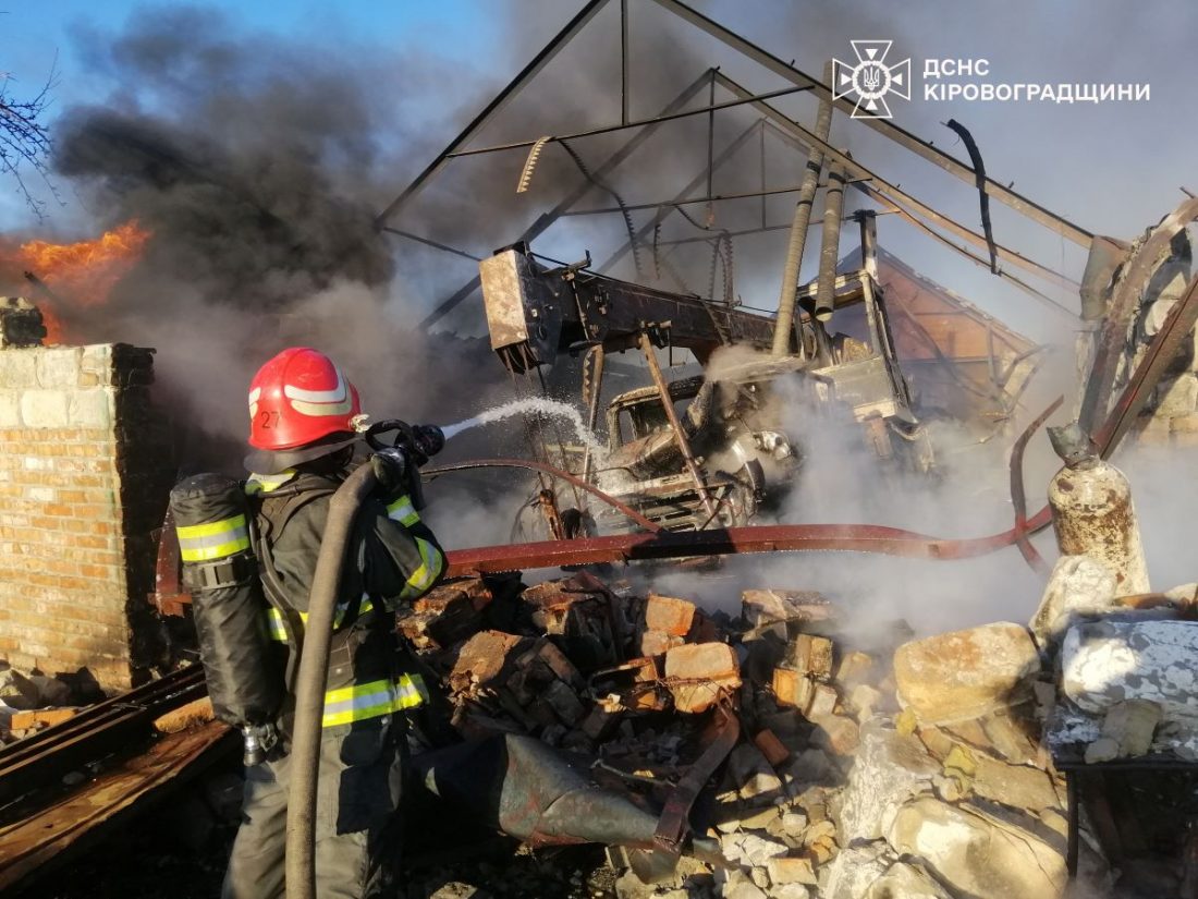 На зображенні показаний пожежник у червоному шоломі та спеціальному костюмі, який гасить пожежу за допомогою шланга з водою. Навколо нього видно уламки будівлі, дим та вогонь. На задньому плані конструкції виглядають пошкодженими від пожежі. На зображенні також є емблема ДСНС Кіровоградщини.