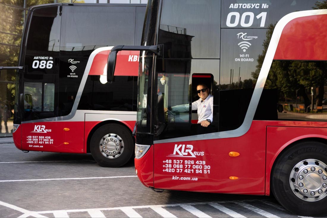 Два двоповерхові червоні автобуси компанії KLR Bus, стоять на парковці. На автобуса написано номер 086 і 001. У вітрині одного з автобусів видно водія в сонцезахисних окулярах, який сидить за кермом.
